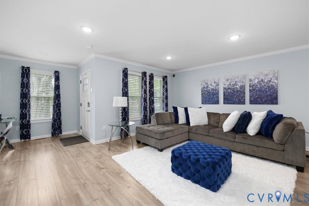 Recessed lighting illuminates the open living space, featuring wood-finish flooring, white crown molding, and multiple windows with blinds
