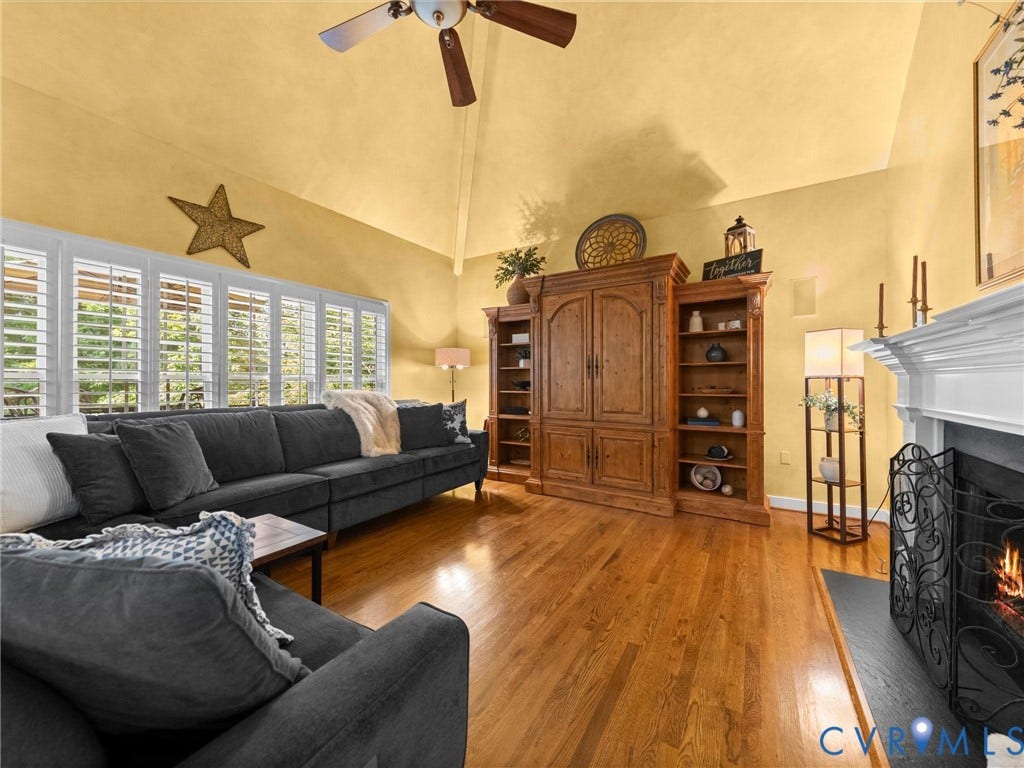 15706 Fox Marsh Drive, Moseley, VA 23120 Vaulted ceilings with a ceiling fan, hardwood flooring, multiple windows with white shutters, built-in wooden shelving, and a fireplace with a white mantel and black hearth