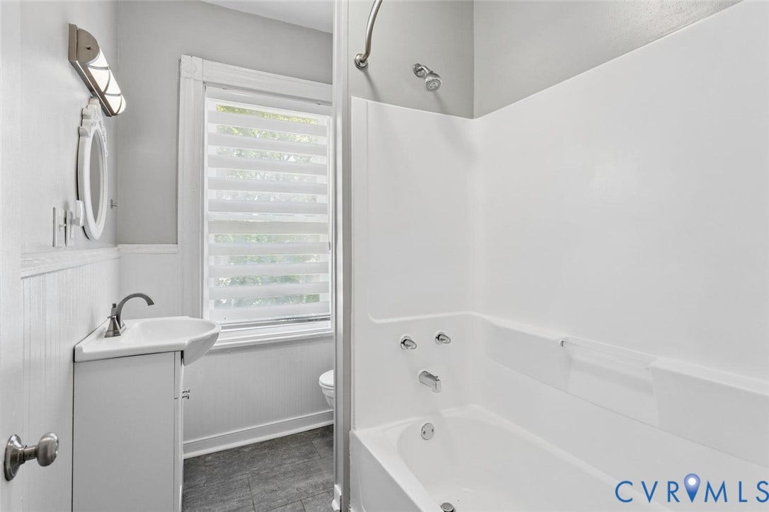 313 W 27th Street, Richmond, VA 23225 White bathtub with integrated shower, white vanity with corner sink, and a modern wall-mounted light fixture