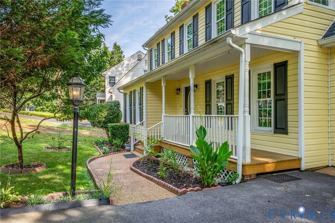 4820 Cedar Cliff Road, Chester, VA 23831 Charming Colonial-style residence featuring soft yellow exterior siding with black-shuttered windows, a welcoming front porch with white railings and columns, and manicured landscaping including mature trees, flowering shrubs, and mulched garden beds