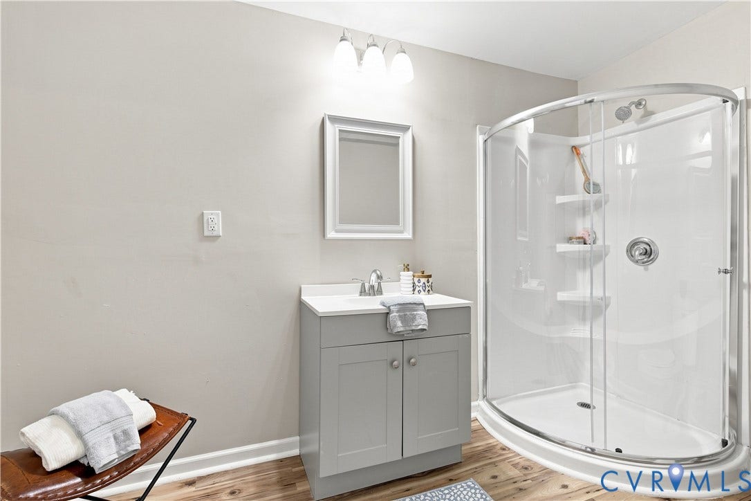 16111 Walkers Mill Road, Stony Creek, VA 23832 Wood-finish flooring, single vanity with light grey cabinetry, white countertop, framed mirror, and a curved glass shower enclosure with built-in shelving