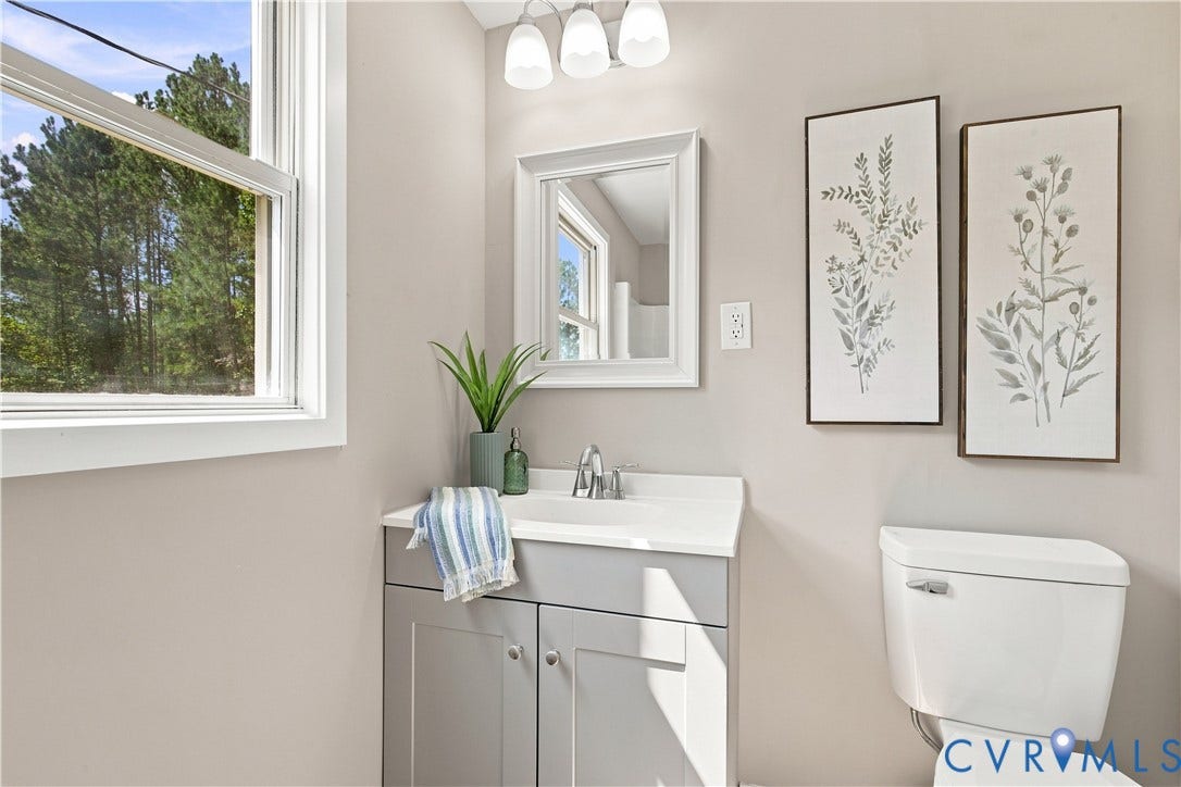 16111 Walkers Mill Road, Stony Creek, VA 23832 White-framed window, gray vanity with integrated sink, chrome-finish faucet, wall-mounted mirror, and a white toilet