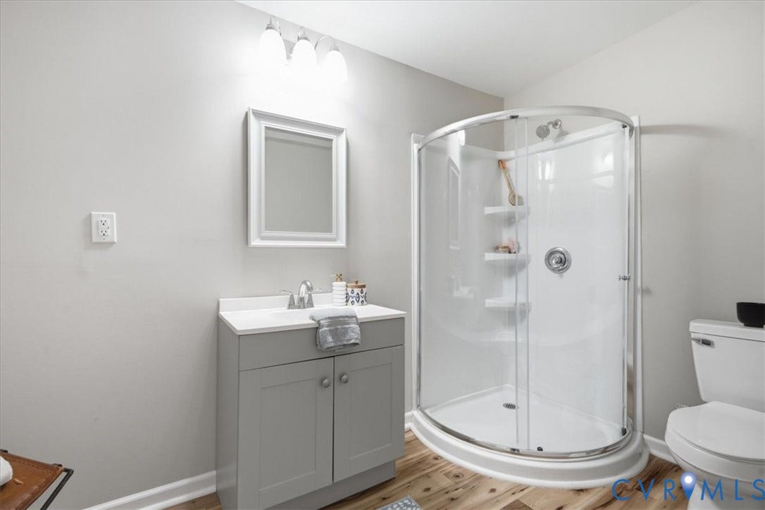 16111 Walkers Mill Road, Stony Creek, VA 23832 Modern bathroom featuring a curved glass shower enclosure with chrome fixtures, gray vanity cabinetry with integrated sink and countertop, framed mirror with integrated lighting, wood-finish flooring, and white ceramic toilet