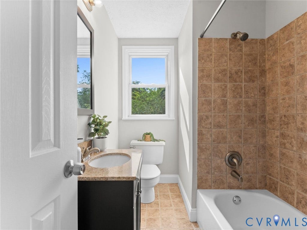 4818 Castlewood Road, Richmond, VA 23234 Renovated bathroom featuring a black vanity with integrated sink and quartz countertop, ceramic tile surround in warm earth tones, soaking tub with shower enclosure, toilet, and natural light from a casement window overlooking landscaped grounds