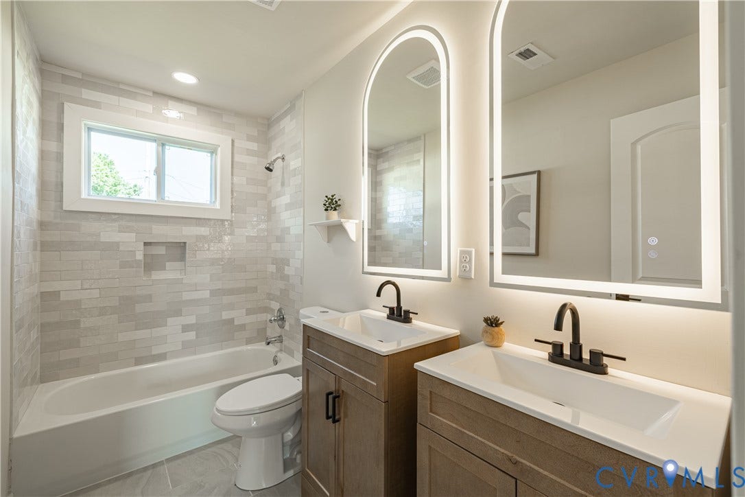 Spa-inspired primary bathroom featuring dual wood-finish vanities with white quartz countertops, matte black faucetry, and illuminated arched mirrors