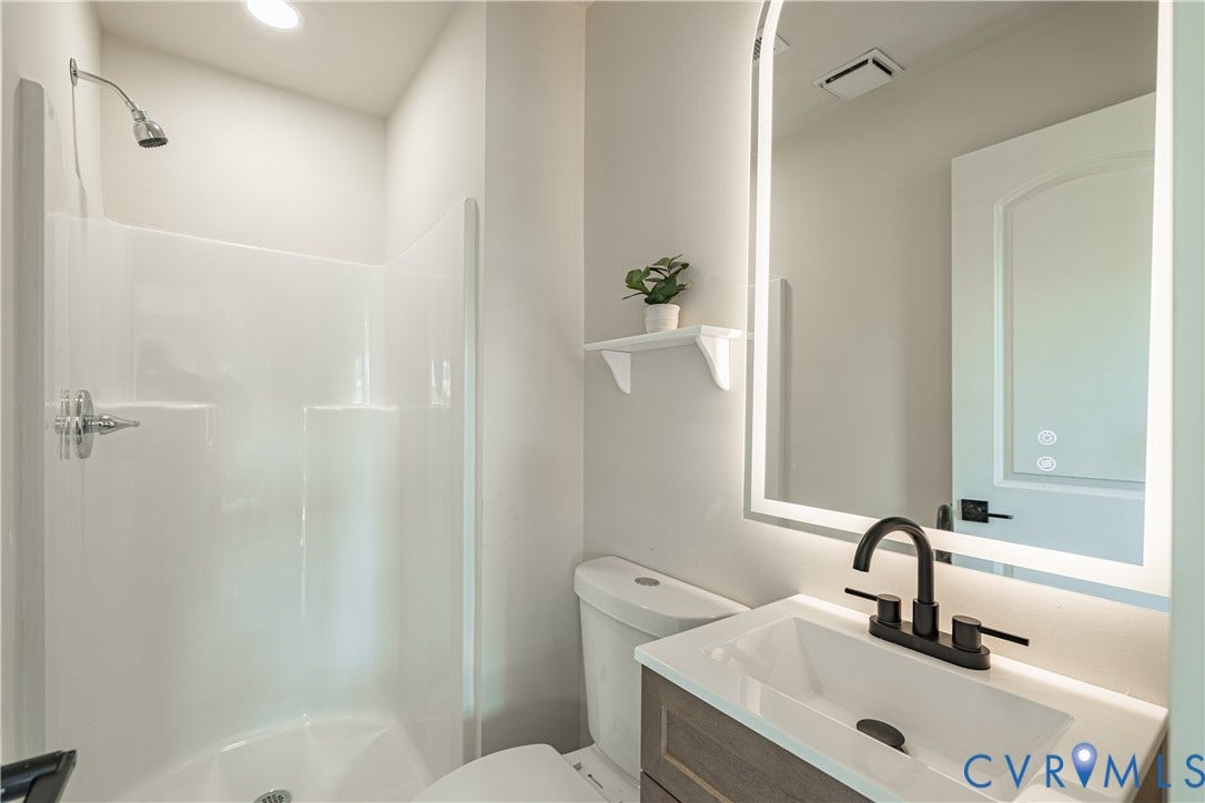 White shower stall with chrome fixtures, vanity with a white countertop and rectangular vessel sink, matte black faucet, and an illuminated mirror