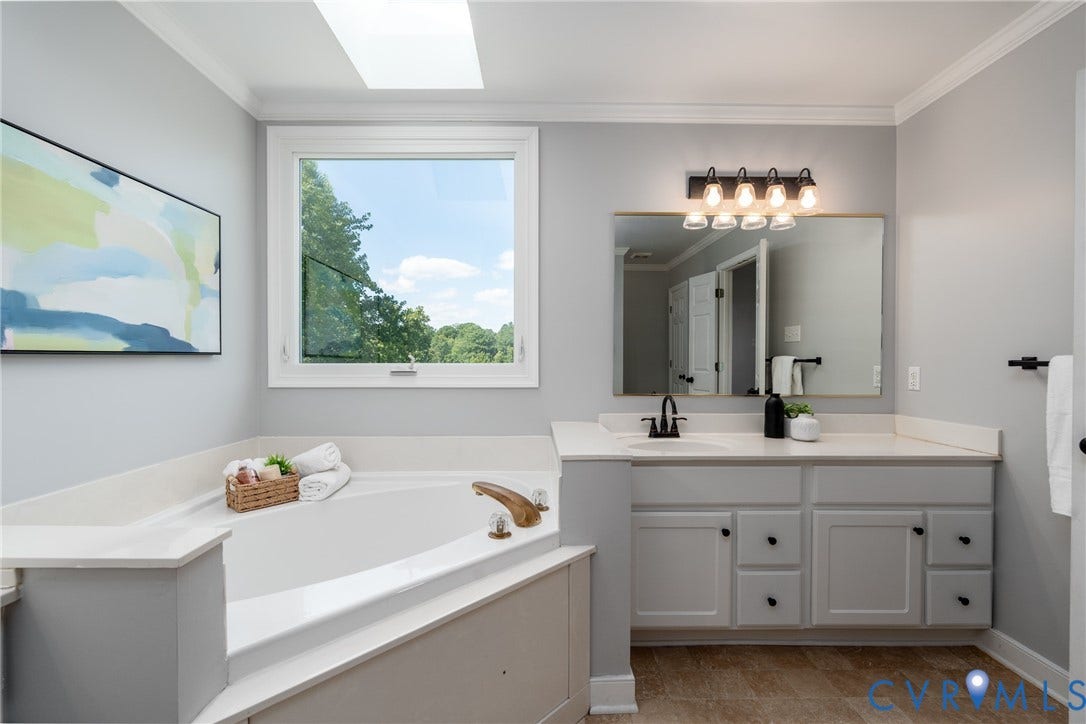 Soaking tub with deck surround, skylight, window, vanity with integrated sink, and wood-finish flooring