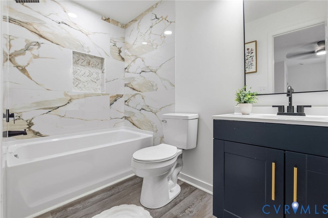 409 S Jefferson Street, Bolling, Petersburg, VA 23803 Spa-inspired bathroom featuring elegant marble-look tile wall surround with integrated niche, white soaking tub, low-profile toilet, and vanity with black cabinetry, brushed gold hardware, and rectangular vessel sink