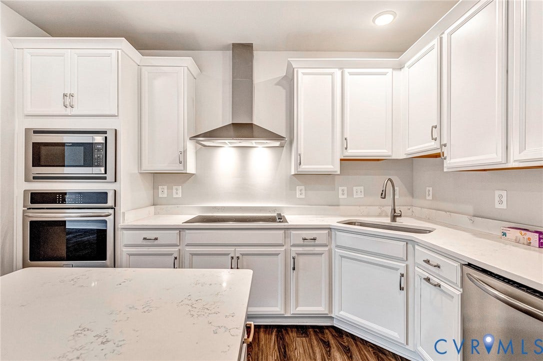 4347 Saunders Station Loop, #A, Glen Allen, VA 23233 Modern kitchen featuring white cabinetry with brushed nickel hardware, quartz countertop with waterfall edge, stainless steel appliances including dual wall ovens and cooktop, undermount sink with chrome faucet, and a stainless steel range hood with backsplash lighting