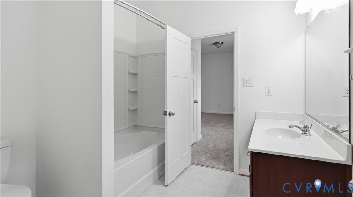 White bathtub with integrated shelving, dark wood-finish vanity with white countertop, and wall-mounted mirror