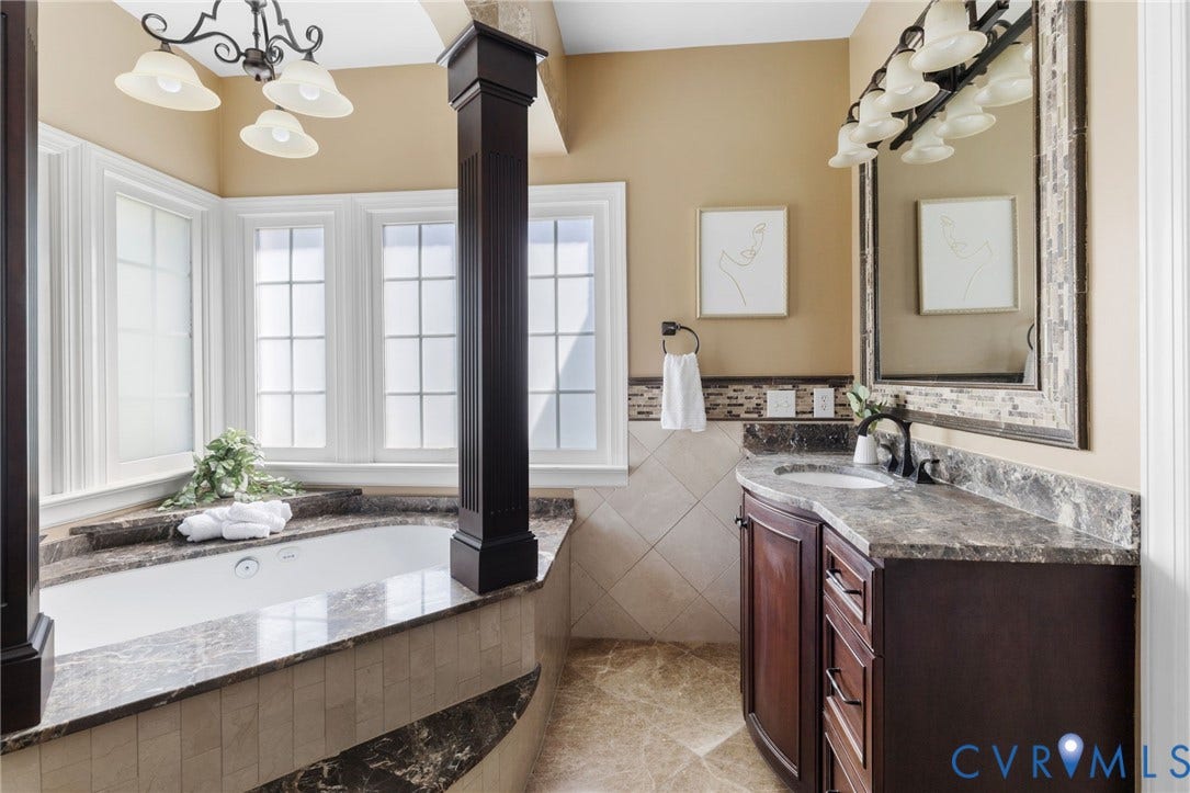2365 Manakintown Ferry Road, Powhatan, VA 23113 Corner soaking tub with a surrounding tile and stone accent, bay windows with grid pattern, single vanity with stone countertop and undermount sink, dark wood-finish cabinetry, and a large framed mirror