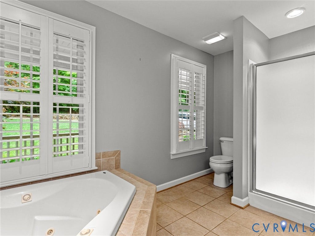 Built-in corner soaking tub, tile flooring, two windows with plantation shutters, enclosed shower, and a toilet