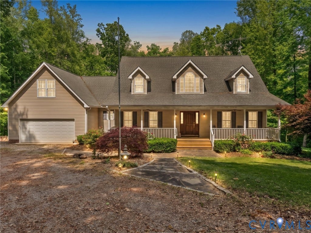 Wood-paneled front door, covered porch with white railing, dormer windows, attached garage with automatic door, and exterior lighting