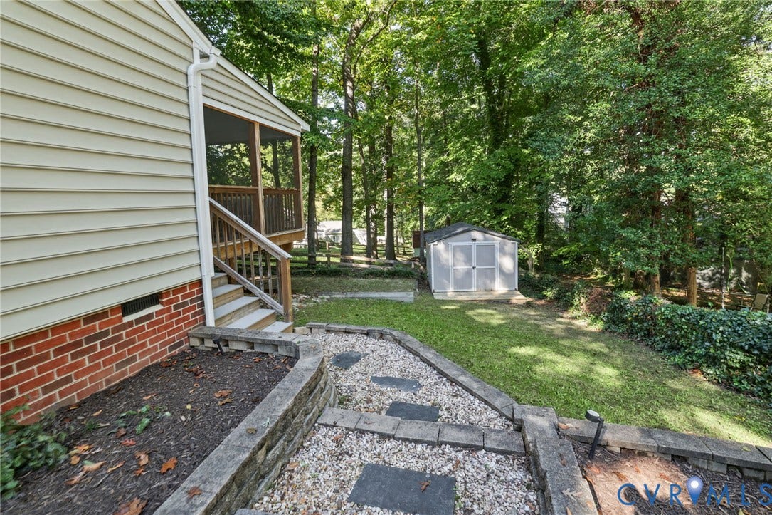 13512 Master Stag Drive, Midlothian, VA 23112 Exterior space featuring a tiered landscape design with stone-block retaining walls, gravel pathways, a screened wooden deck, and a detached storage shed set against a mature woodland backdrop