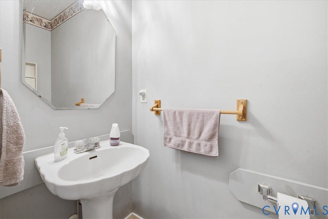 13308 S Chester Road, Chester, VA 23831 Elegant powder room featuring a white pedestal sink, octagonal framed mirror with decorative molding, brass towel bar hardware, and crisp white wall finishes with coordinating tile wainscoting