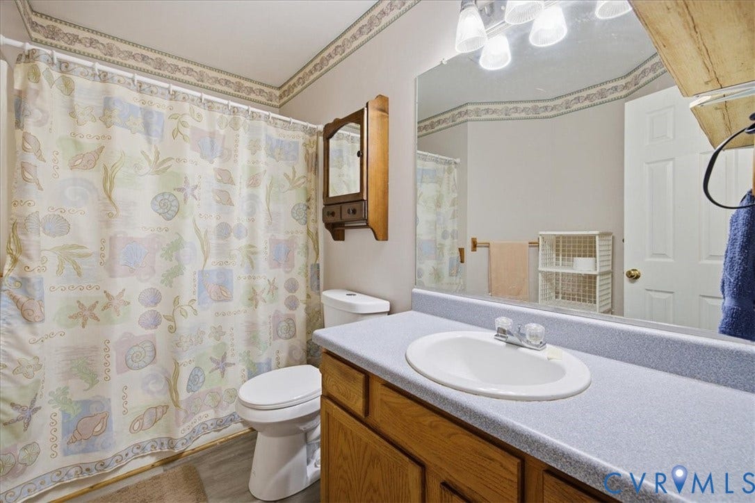 13308 S Chester Road, Chester, VA 23831 Bright, well-appointed bathroom featuring a wood-finish vanity with dual-basin sink, gray quartz countertop, large framed mirror with integrated lighting, and patterned shower curtain