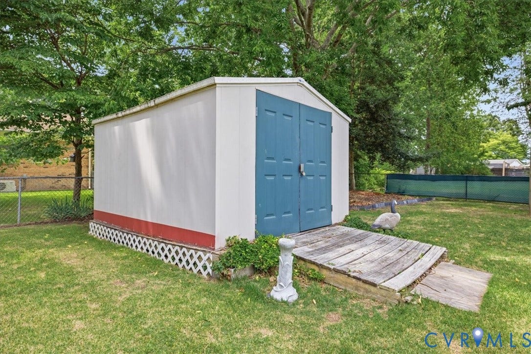 13308 S Chester Road, Chester, VA 23831 Charming storage shed featuring a blue double-door entrance, white exterior siding with contrasting red trim, lattice foundation detailing, and a wooden ramp for accessibility