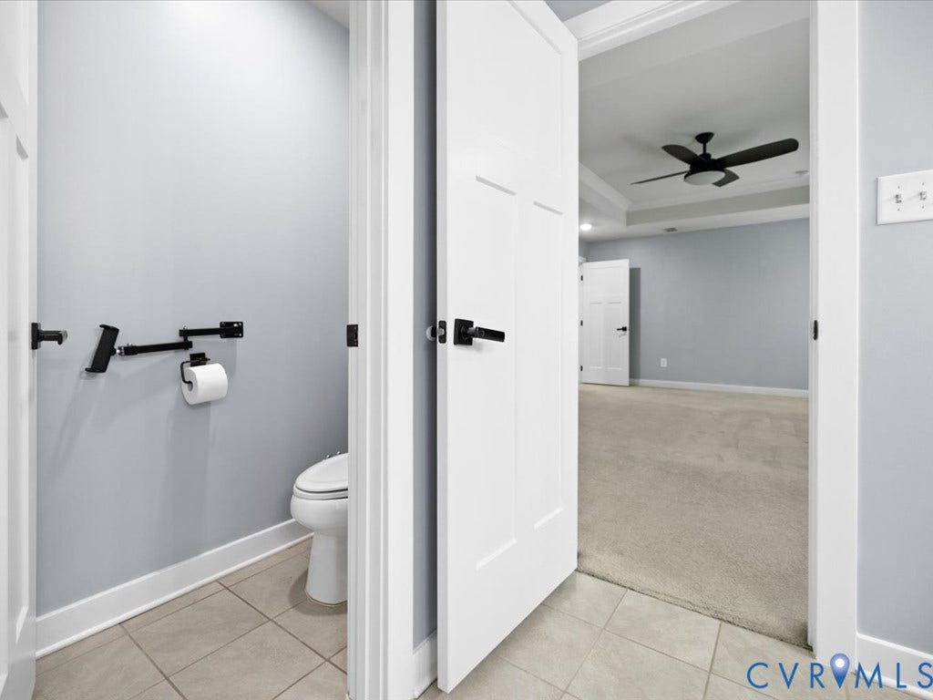 Powder room featuring a toilet and tile flooring, with an adjoining room showcasing carpet flooring, a tray ceiling, and a ceiling fan