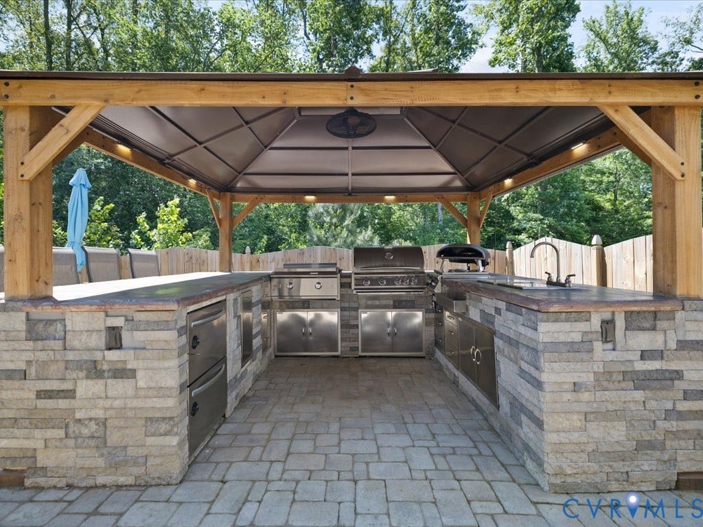 Outdoor kitchen featuring a wood-frame gazebo, built-in stainless steel appliances, integrated sink with faucet, stone-clad island, and paver flooring