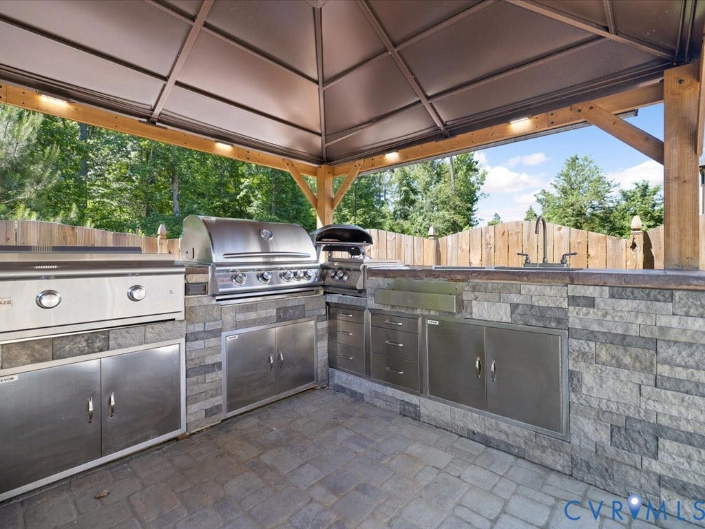Outdoor kitchen featuring a stone veneer island, stainless steel appliances, a built-in sink, and a paved stone surface, all under a permanent gazebo structure