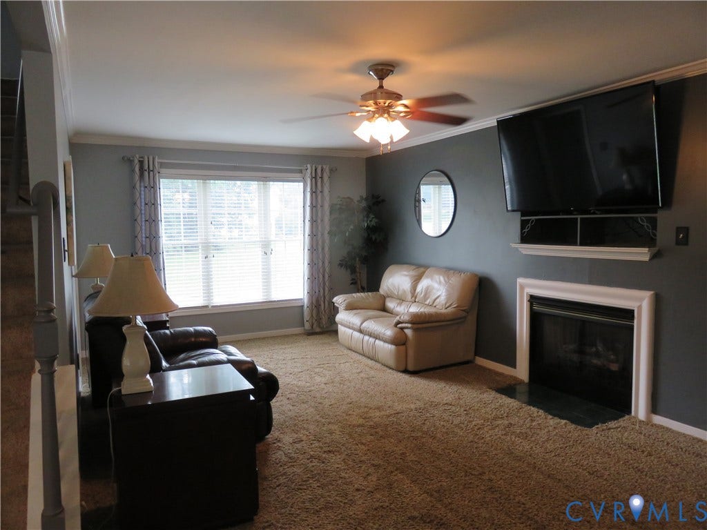 3107 McCabe Court, Chester, VA 23831 Carpeted room with a ceiling fan, a prominent window, and a built-in fireplace featuring a white surround and a dark hearth