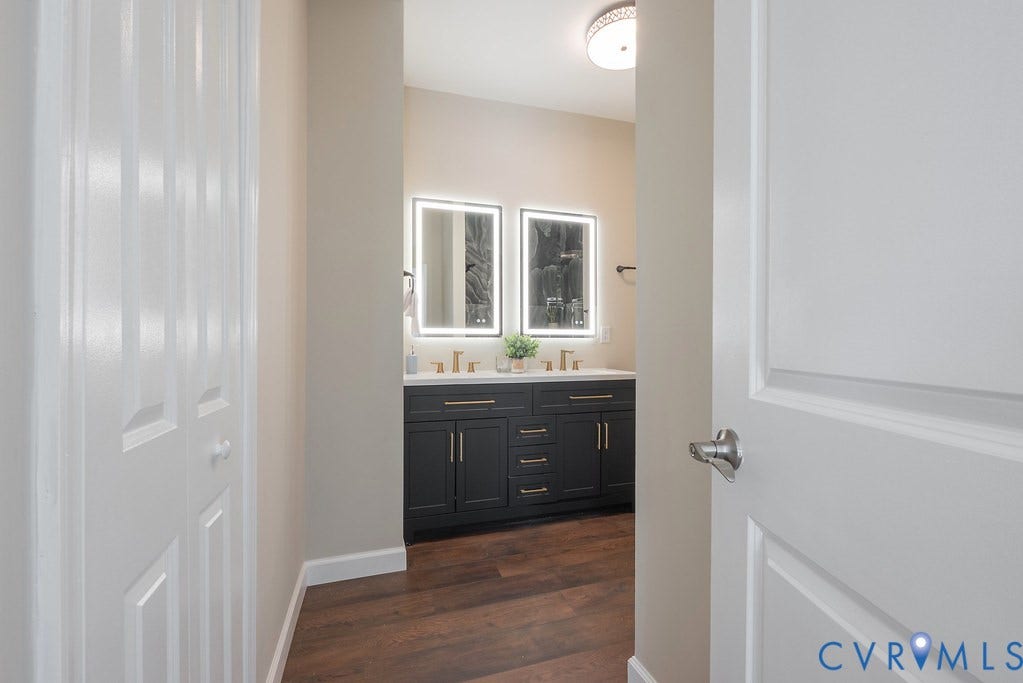 Bathroom featuring a dual vanity with dark cabinetry, gold-finish hardware, two illuminated mirrors, and wood-finish flooring