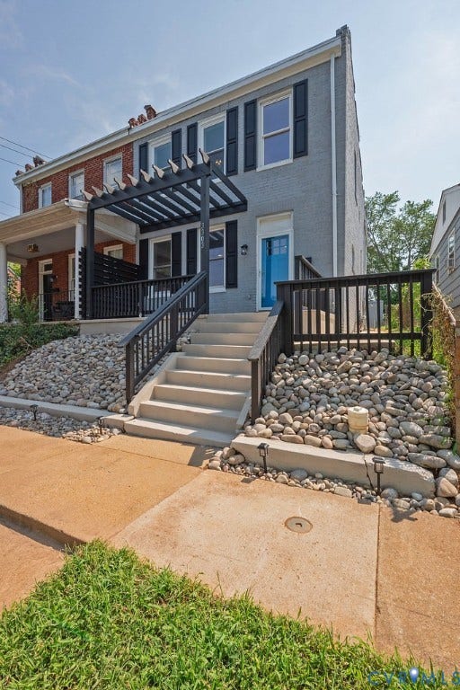 Two-story residence featuring a pergola-covered porch with black railings, a bright blue front door, and a concrete pathway leading to the entrance
