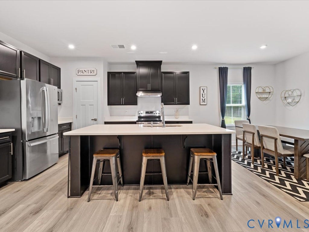 Wood-finish flooring throughout, dark cabinetry, stainless steel appliances, kitchen island with seating, and a dedicated pantry