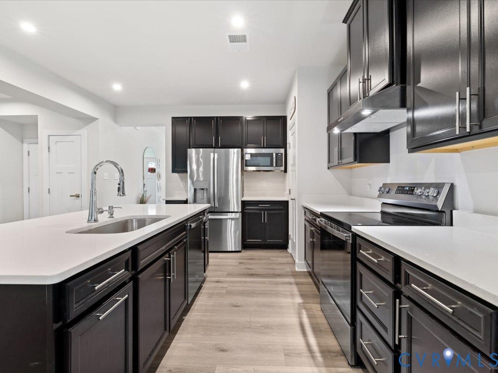 Modern kitchen featuring a generous island with white quartz countertop and waterfall edge, stainless steel appliances including a built-in microwave, dark cabinetry with brushed metal hardware, white subway tile backsplash, and wood-finish flooring throughout the open layout