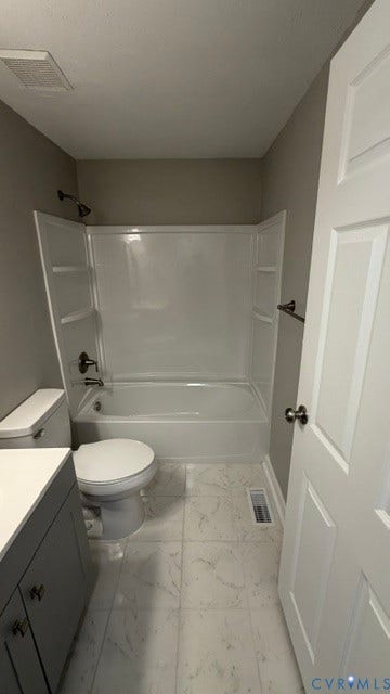 White bathtub and shower surround with dark bronze fixtures, gray vanity with white countertop, toilet, and white patterned floor tiles
