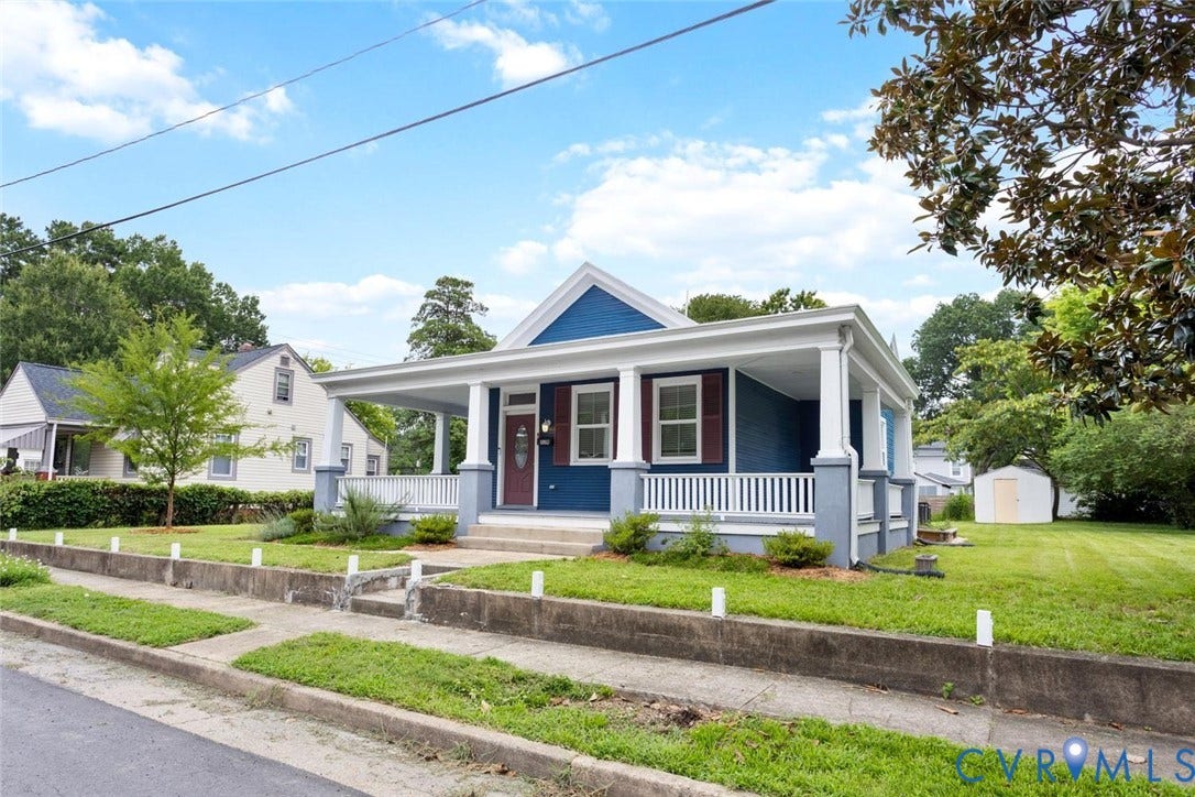 Blue facade with white trim, covered front porch, red accent shutters, and a front yard with mature landscaping