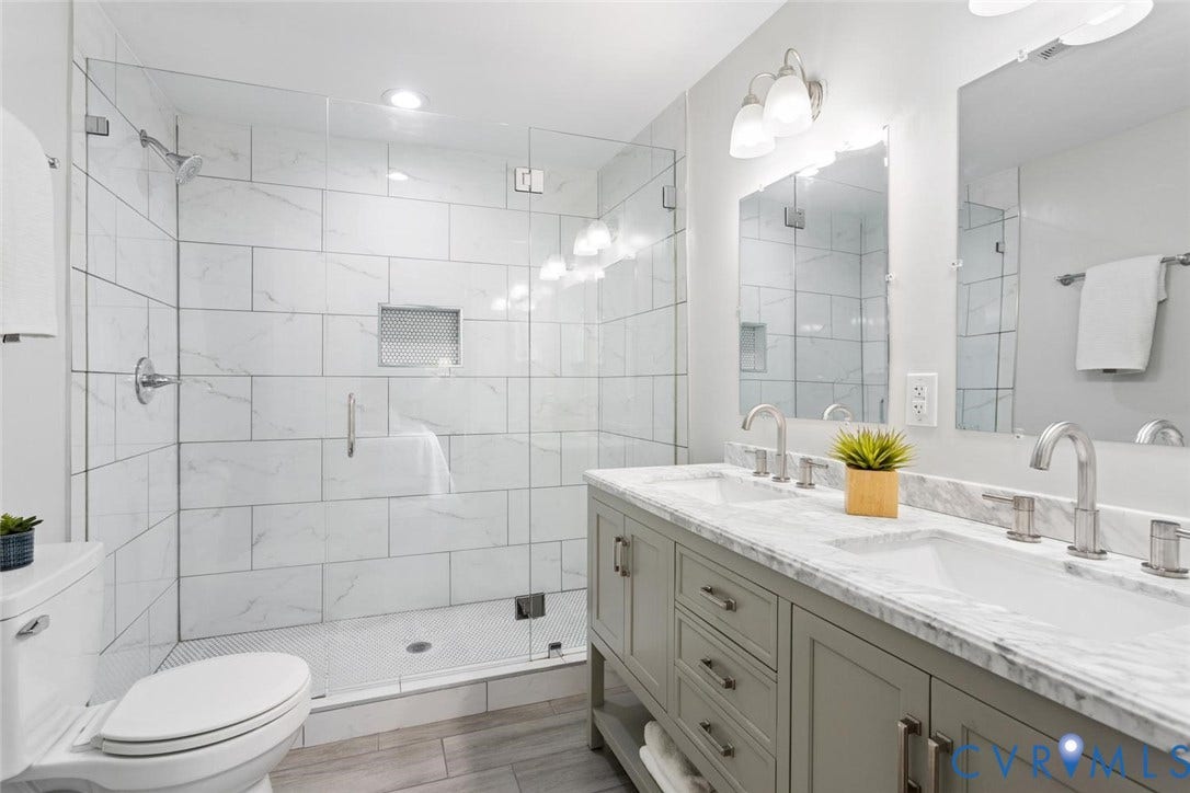 Dual sink vanity with stone countertop, walk-in shower with glass enclosure, white tiled shower surround, recessed shower niche, and wood-finish flooring