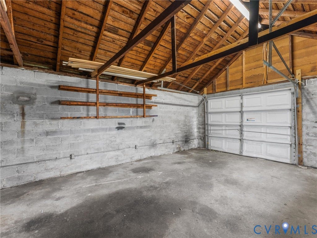 Spacious garage featuring a concrete floor, exposed wood beam ceiling, block walls, built-in shelving, and a panel garage door