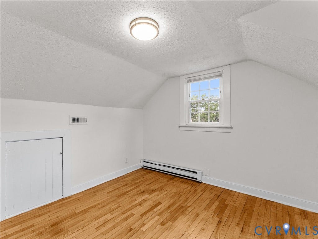 Room featuring wood flooring, a window providing natural light, white painted walls, a ceiling-mounted light fixture, and a baseboard heating unit