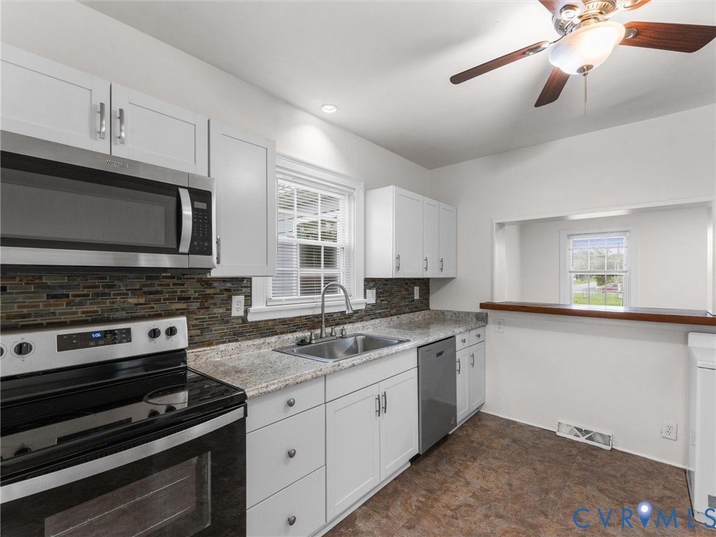 Updated kitchen featuring white shaker cabinetry, stainless steel appliances, a multi-tone tile backsplash, light-colored countertops, and a pass-through window with a wood-finish bar