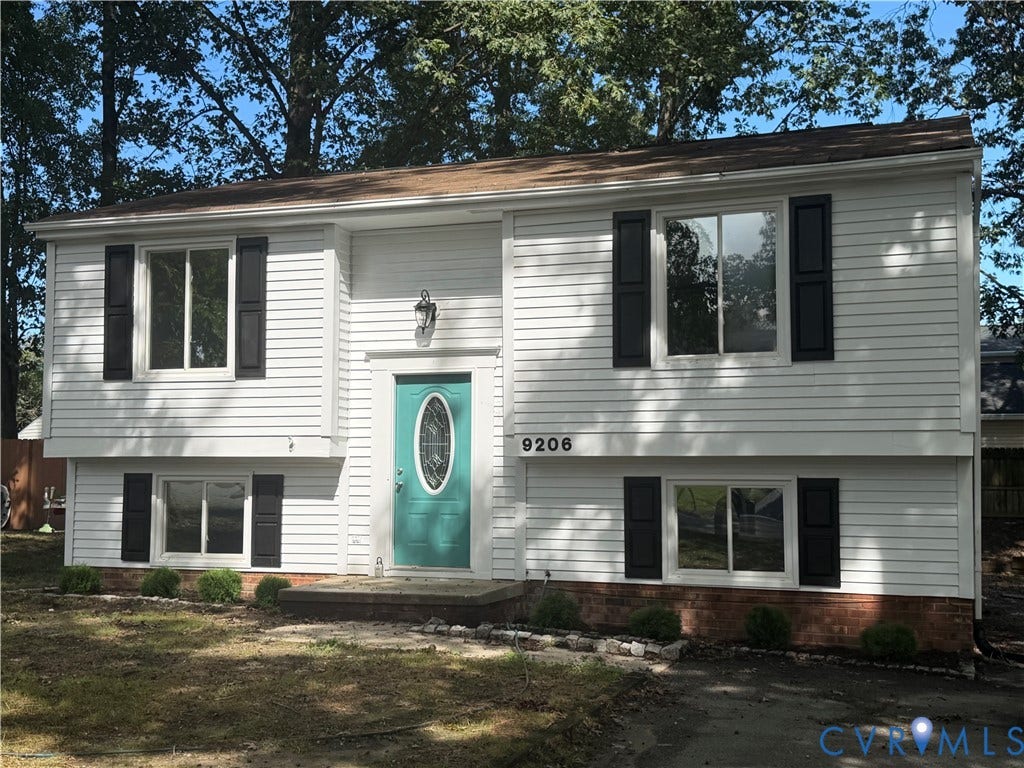 White siding exterior with black shutters, a teal entry door with an oval glass insert, and a brick foundation accent