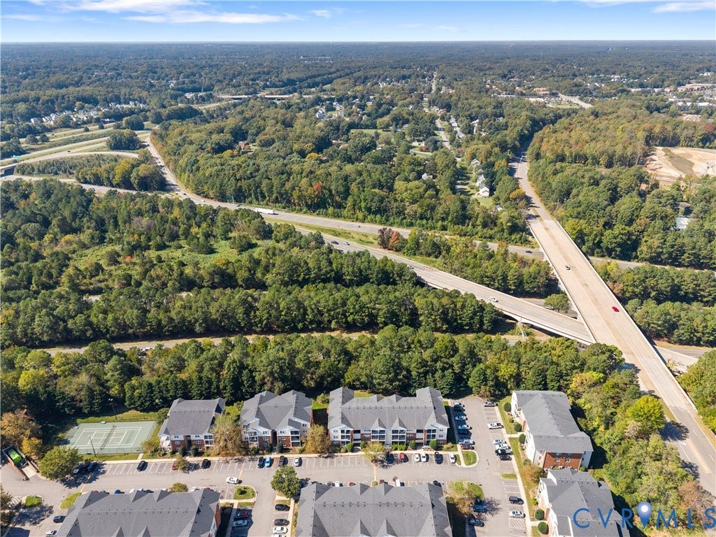 821 Brassie Lane, #E, The Crossings Golf Course Area, Glen Allen, VA 23059 Aerial perspective of a multi-unit residential complex featuring gabled roofs, asphalt parking surfaces, a fenced tennis court, and surrounding mature woodland