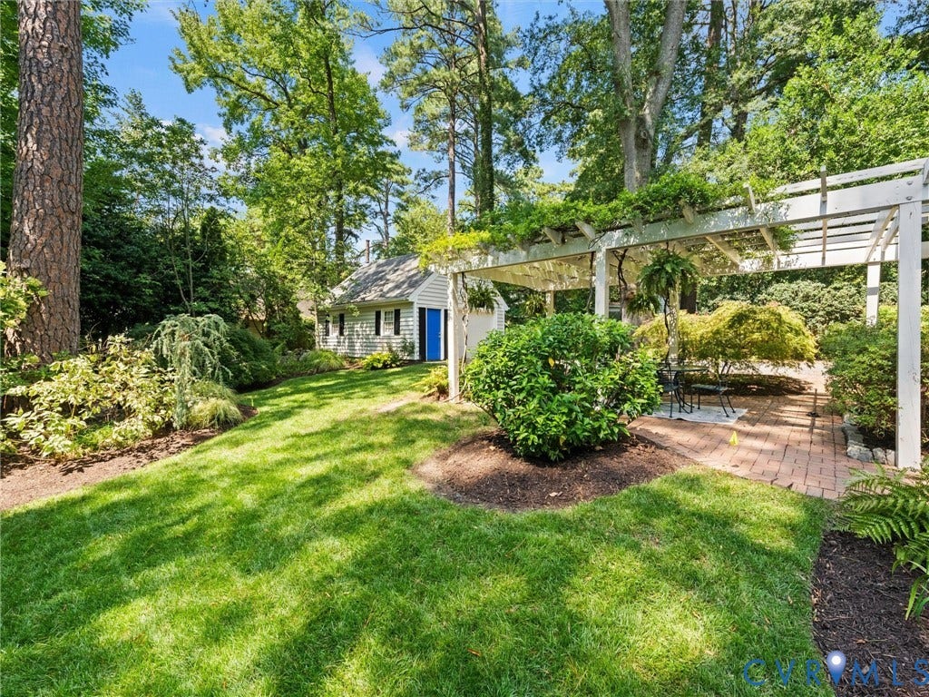 Expansive outdoor space featuring a lush lawn, mature trees, a white pergola, a brick patio, and dense perimeter landscaping