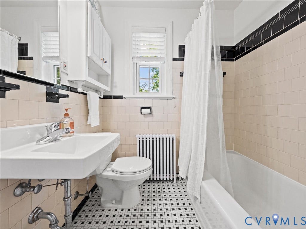 White pedestal sink with chrome faucet, black and white mosaic tile flooring, white tiled walls, and a built-in tub with shower