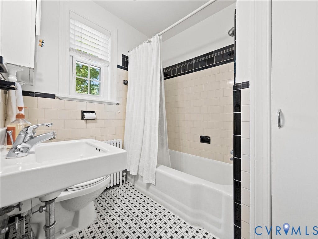 White subway tile shower surround with black accent trim, white bathtub, black and white geometric floor tiling, and a white vanity with a chrome faucet