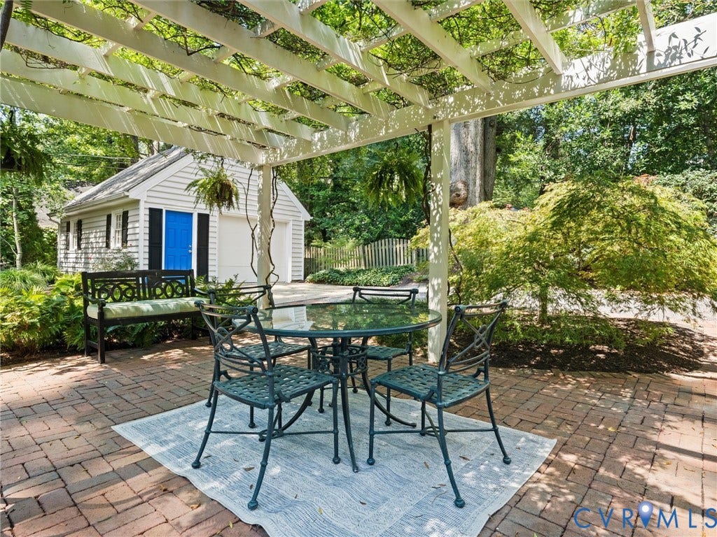 Brick paver patio with a white pergola, mature landscaping, and a detached structure featuring a garage door and blue door