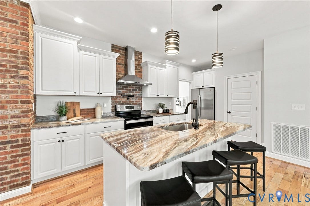 Beautiful kitchen featuring white cabinetry, granite countertops, a large center island with seating, and eye-catching exposed brick accents.