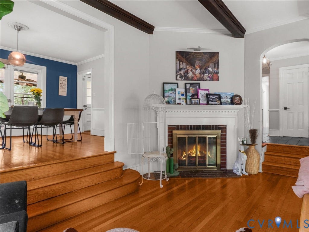 Hardwood flooring throughout, brick fireplace with metal insert, exposed wood ceiling beams, and arched doorway