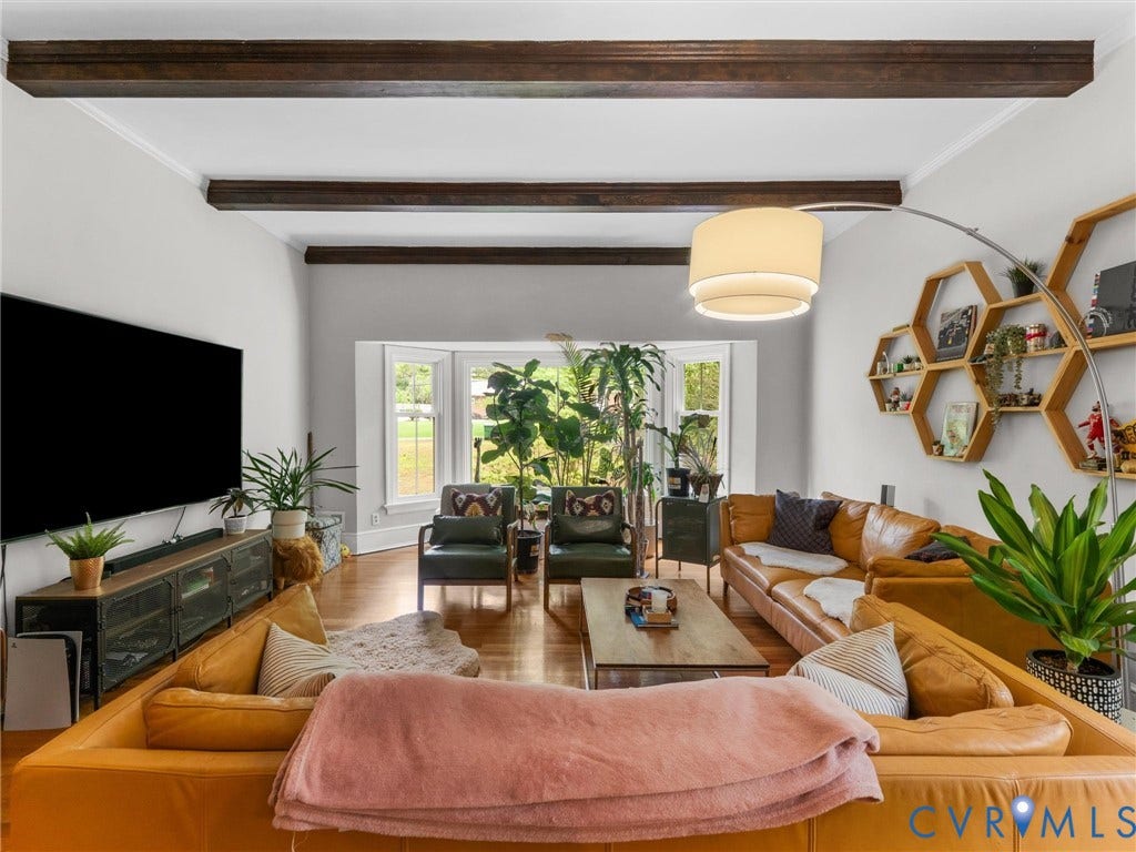 Bright, open-concept living area featuring exposed wood-beam ceiling detail, large multi-pane windows with natural light, wood-finish flooring throughout, and a cylindrical pendant light fixture