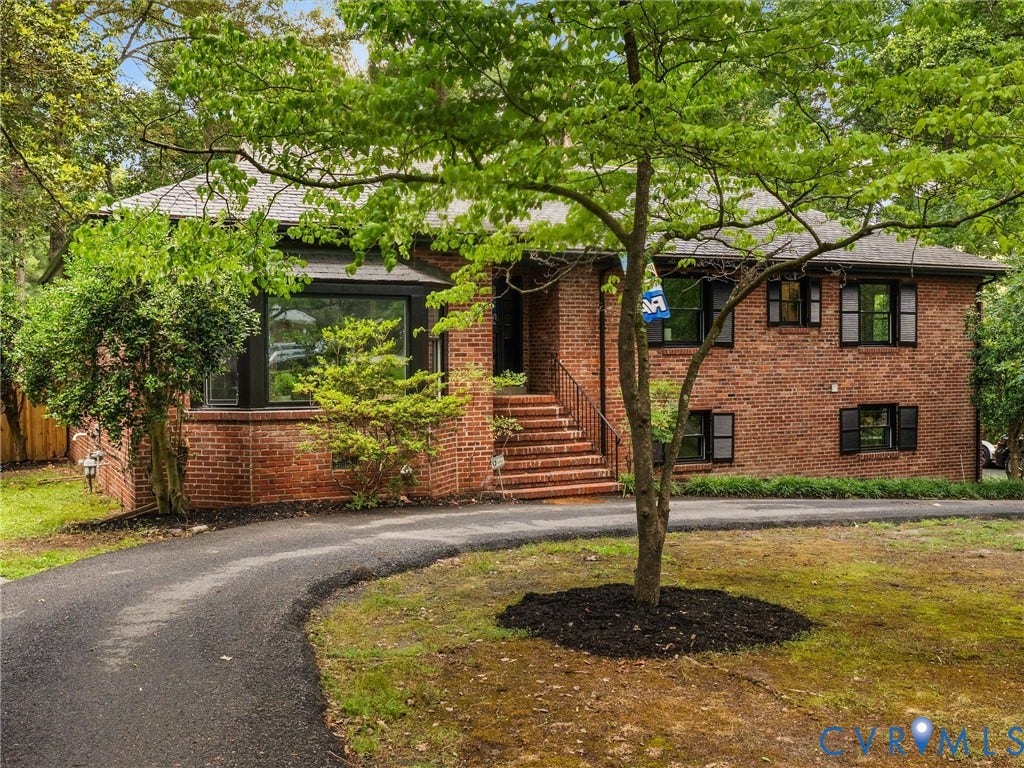 Brick exterior featuring multiple windows with dark shutters, a paved driveway, and mature landscaping