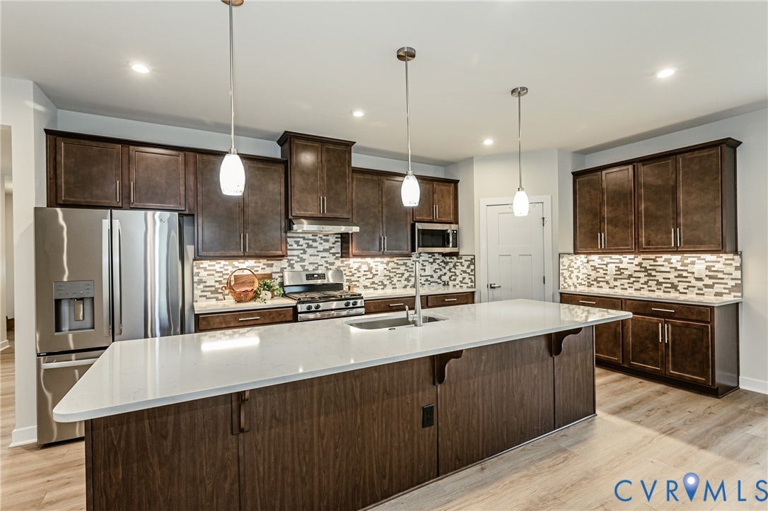 7683 Autumn Leaves Lane, Estates The Groves At New Kent, New Kent, VA 23124 Kitchen featuring a large central island with a light-colored countertop, dark wood-finish cabinetry, stainless steel appliances, and a mosaic tile backsplash