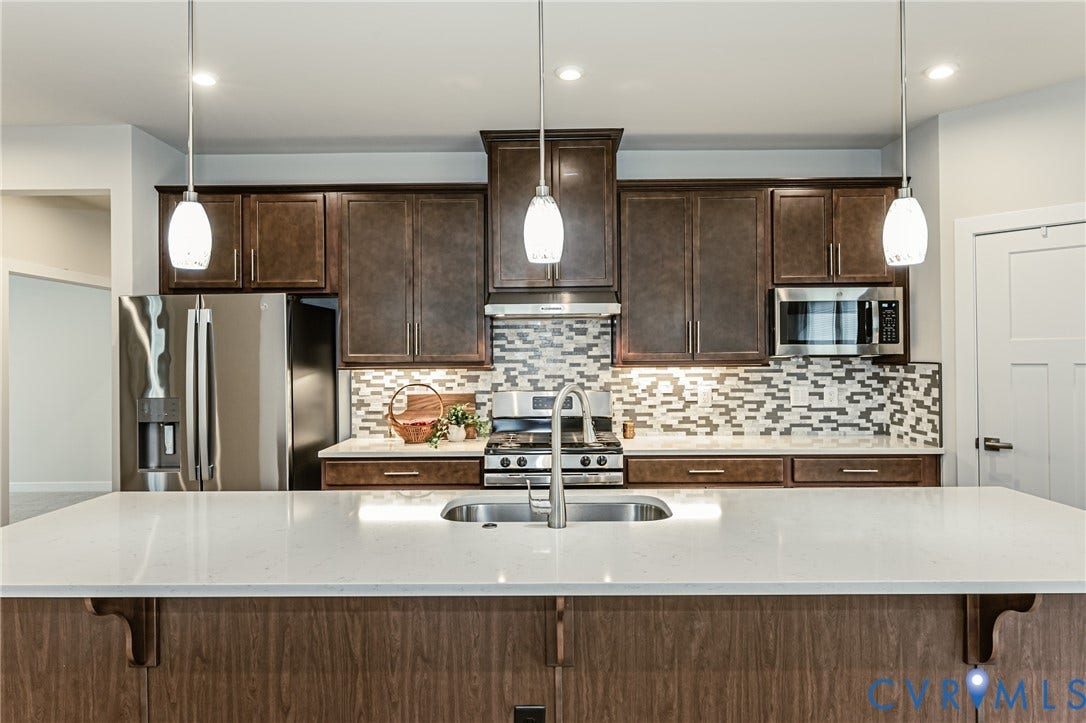 7683 Autumn Leaves Lane, Estates The Groves At New Kent, New Kent, VA 23124 Kitchen featuring dark wood-finish cabinetry, stainless steel appliances, a mosaic tile backsplash, and a large central island with a built-in sink and white countertop