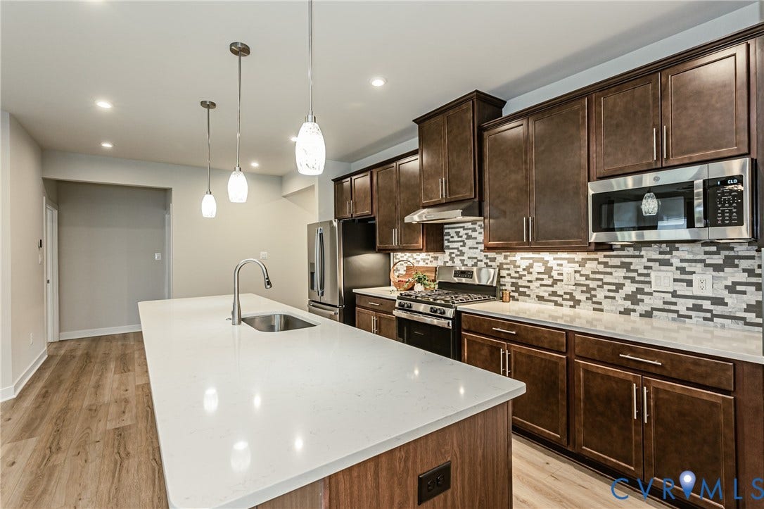 7683 Autumn Leaves Lane, Estates The Groves At New Kent, New Kent, VA 23124 Kitchen featuring a large island with sink, dark wood-finish cabinetry, stainless steel appliances, a mosaic tile backsplash, and recessed lighting