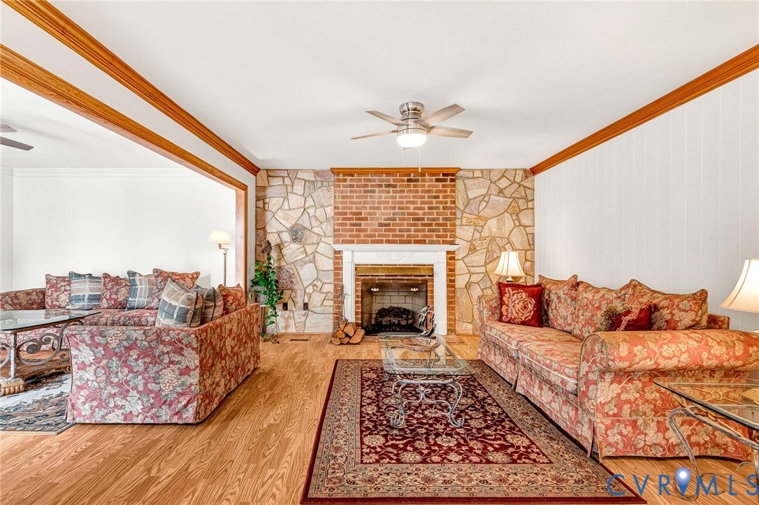 Hardwood flooring, stone and brick fireplace with mantle, exposed wood beams, white wall paneling, and a ceiling fan