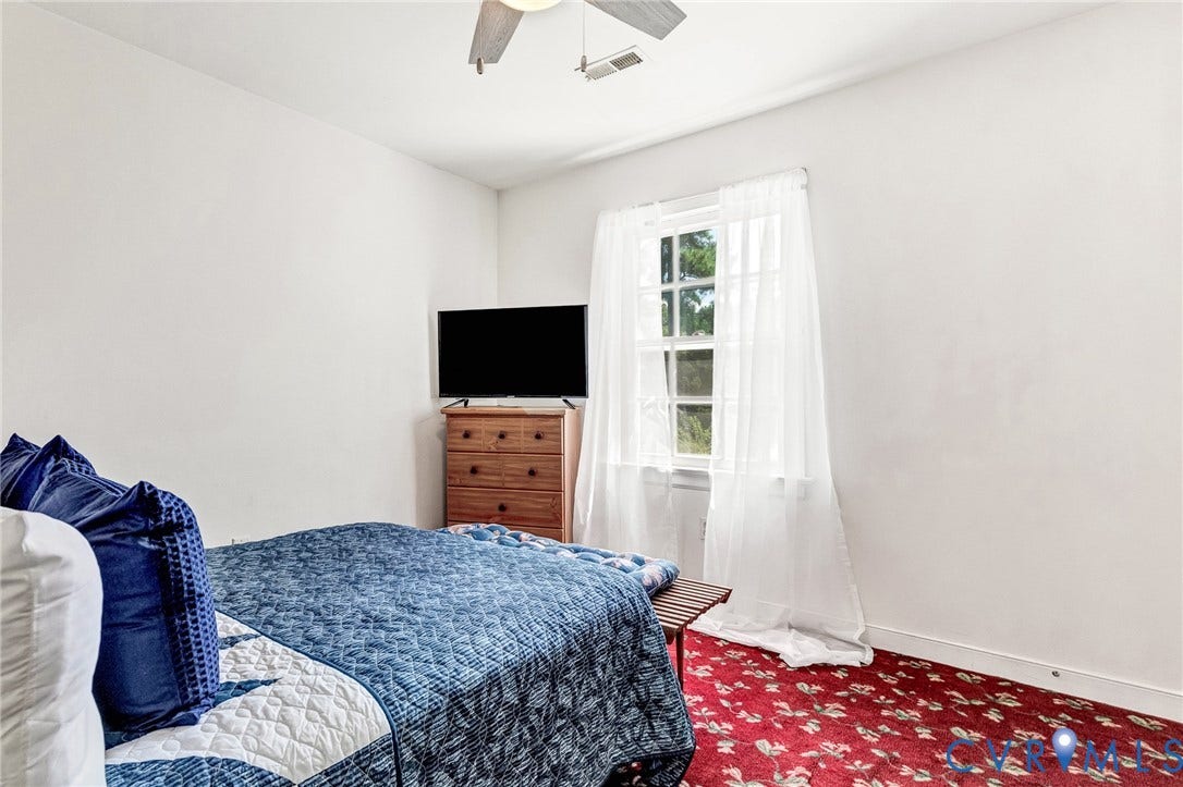 Bright secondary bedroom with natural light from French doors, wood-finish dresser with integrated flat-screen television, ceiling fan, and patterned area rug accenting the hardwood flooring